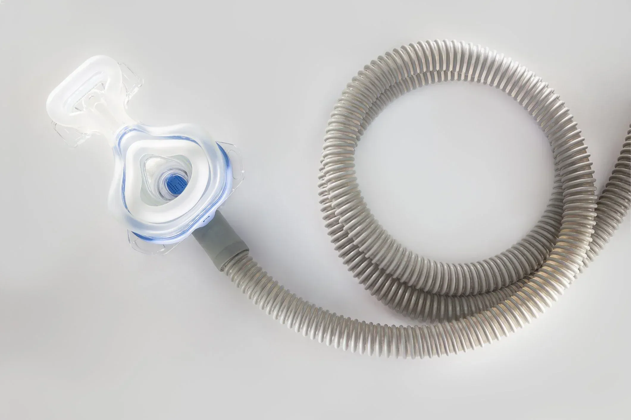 Clear CPAP nasal mask lying beside its neatly coiled grey air hose on a plain white surface