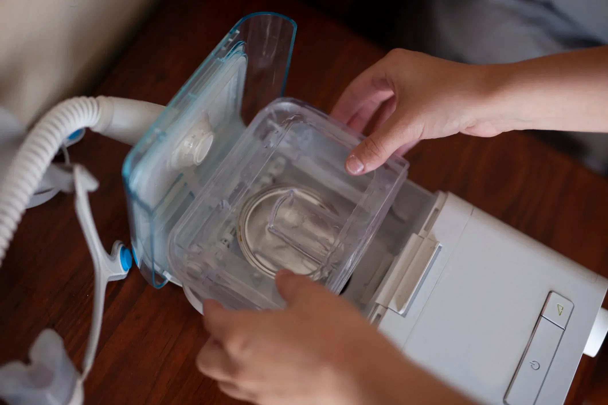 Two hands lifting the clear water chamber out of a CPAP machine on a wooden bedside table