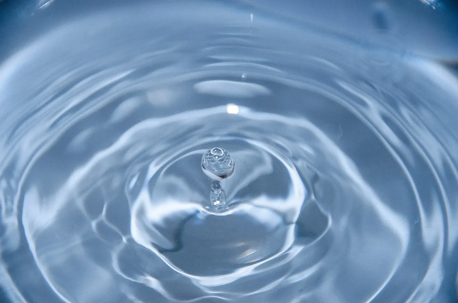 Single water droplet rebounding from a still pale blue surface at the centre of spreading ripples